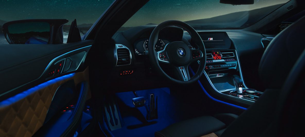 BMW M8 Competition Coupé, Interieur bei Nacht mit M Lederlenkrad und blauem ambienten Licht bei geöffneter Fahrertür.